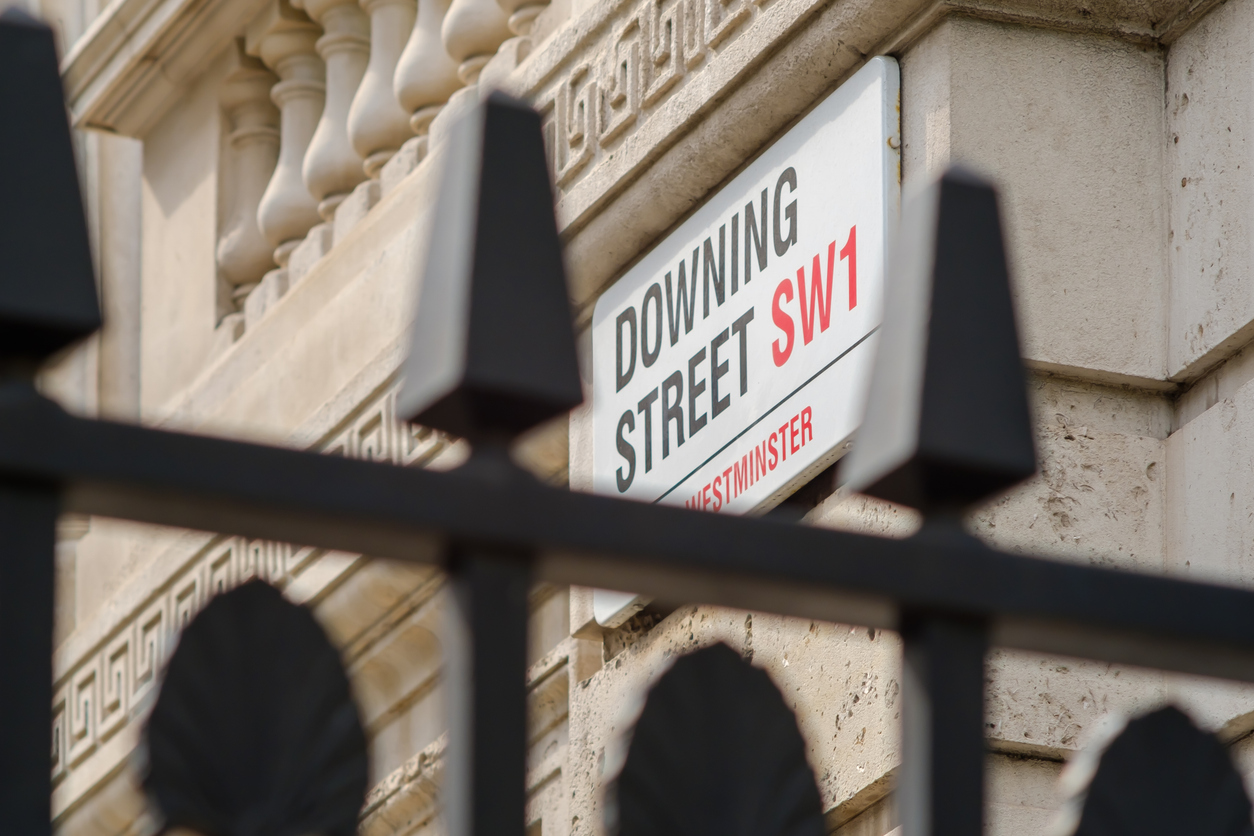 A street sign reading 'Downing Street SW1' is viewed through a black fence